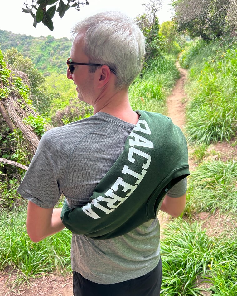 A Seed team member on a dirt trail through green hillside brush, a "Bacteria" sweatshirt slung over one shoulder