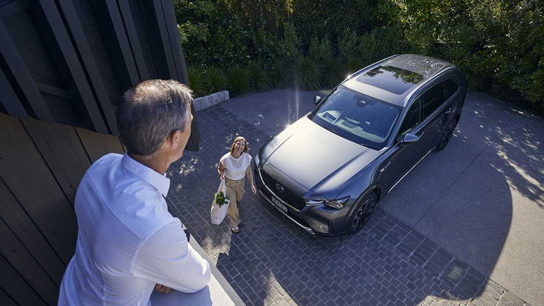 Man on a balcony looking down at his wife exiting her grey Mazda CX-90 SUV in their driveway