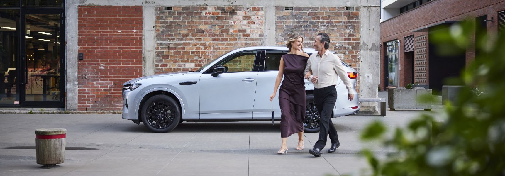 Woman and man walking away from their white Mazda CX-60 SUV that is parked in front of a brick wall 