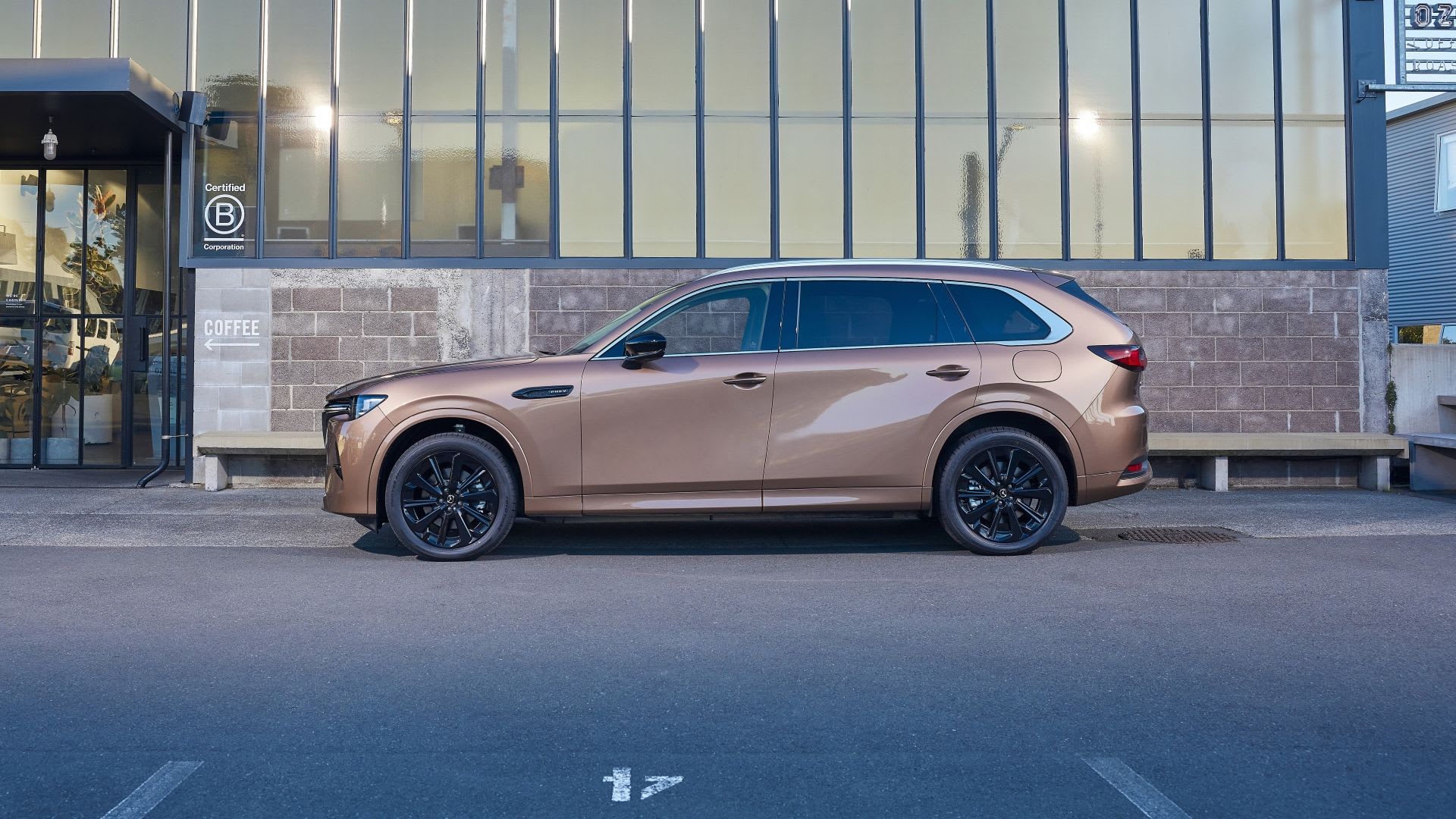 Side view of a Melting Copper Mazda CX-80 7-seat hybrid SUV parked in front of a modern glass and brick office building
