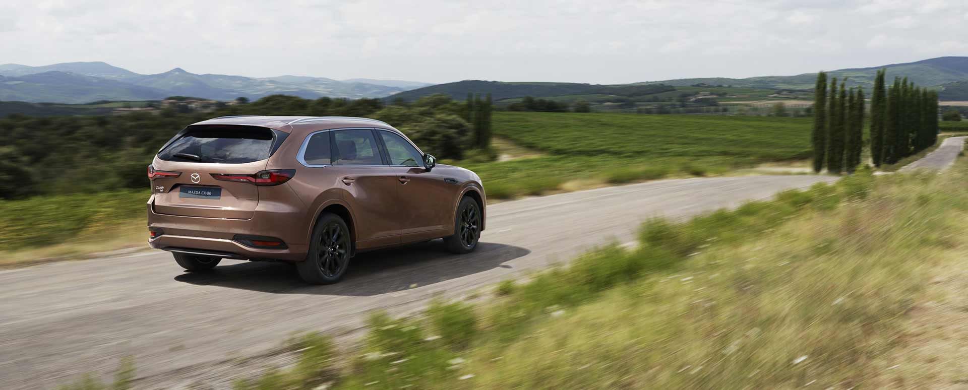 Rear three-quarter view of a Melting Copper Mazda CX-80 7-seat hybrid SUV driving on an open road
