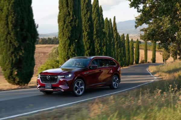 Front three-quarter view of an Artisan Red Mazda CX-80 7-seat hybrid SUV parked on a road lined with pine trees 