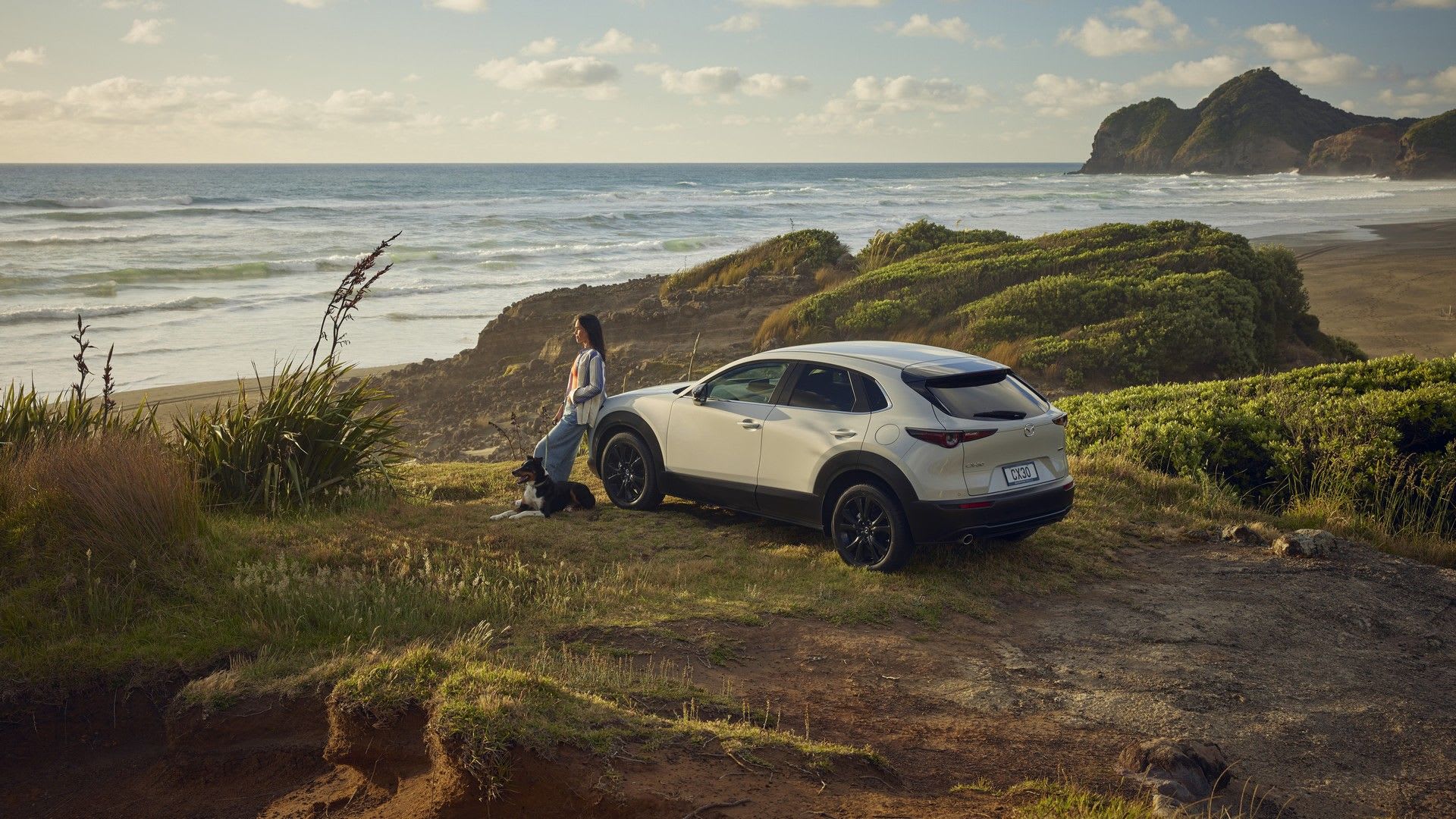 Woman leaning against her white Mazda CX-30 SUV at an ocean lookout