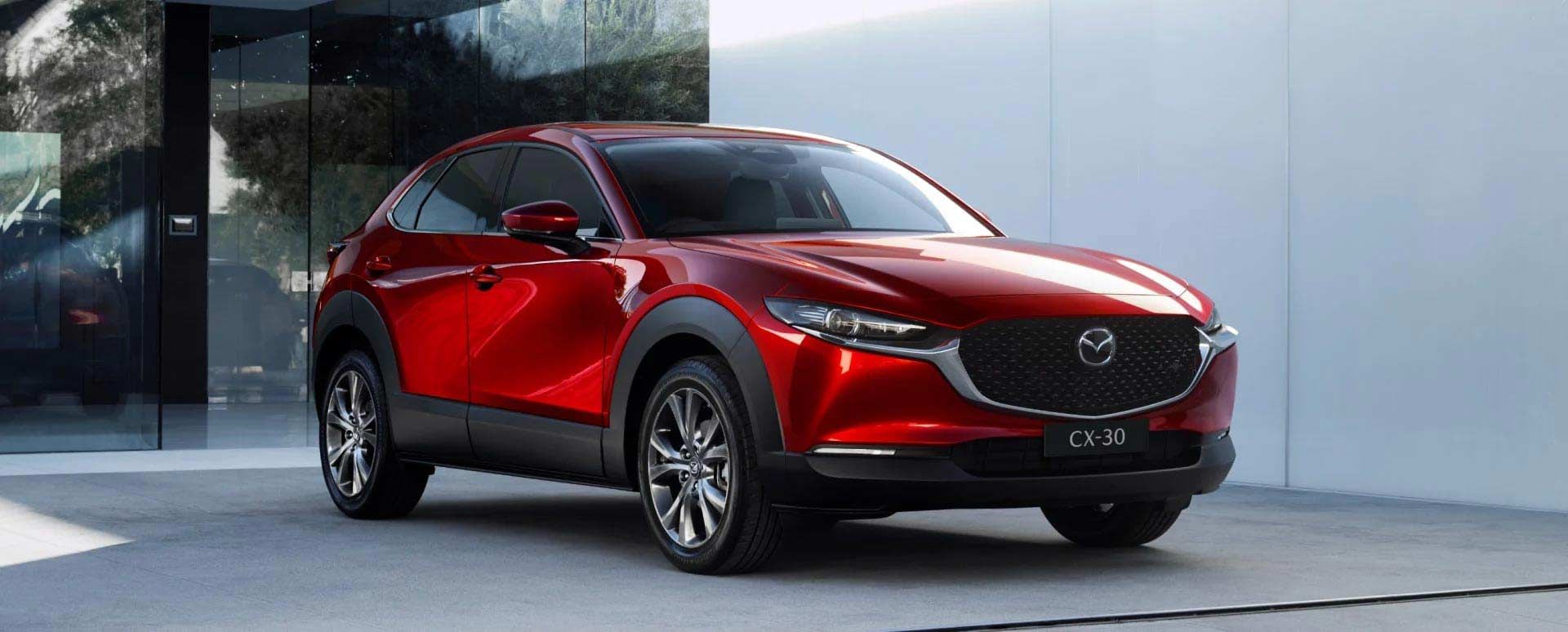 Front three-quarter view of a Soul Red Mazda CX-30 SUV parked in front of a glass and concrete wall