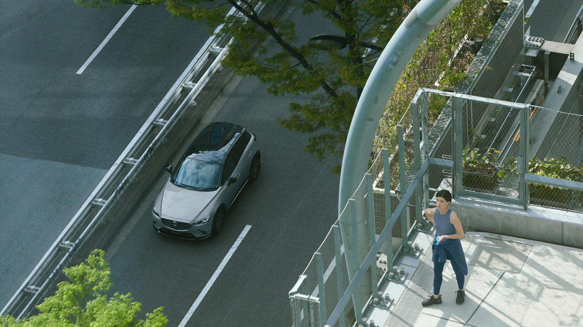 Looking down from a city building onto a silver Mazda CX-3 SUV driving along a 4 lane road