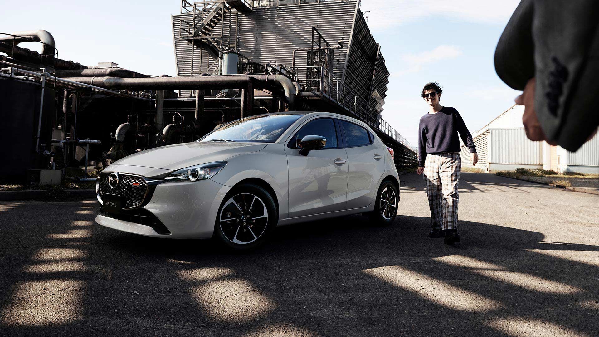 Front three-quarter view of a Snowflake White Mazda2 hatch parked in front of a factory with a man walking towards the car
