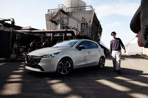 Front three-quarter view of a Snowflake White Mazda2 hatch parked in front of a factory with a man walking towards the car