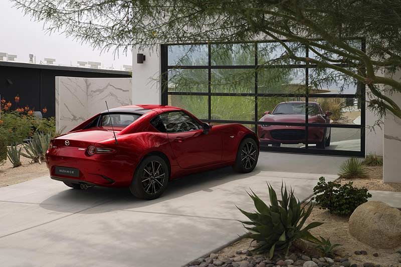 Rear three-quarter view of a Soul Red Mazda MX-5 RF parked on a driveway in front of a modern garage 