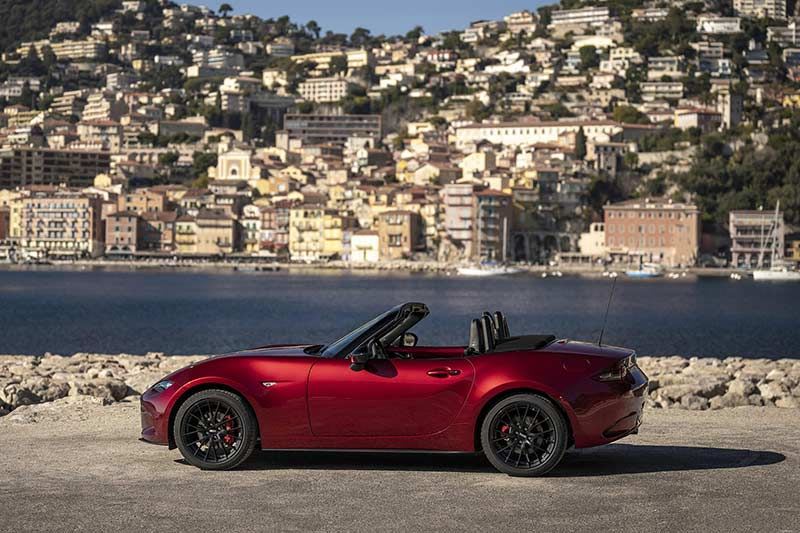 Side view of a Soul Red Mazda MX-5 roadster parked in front of a river and hillside European city