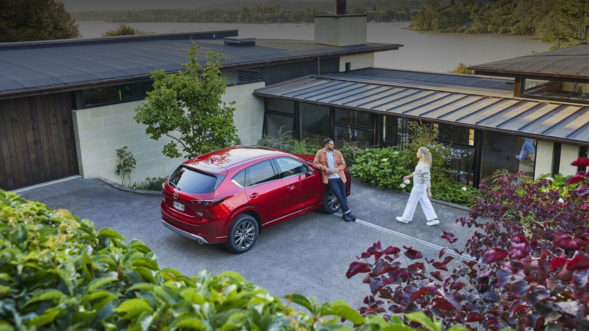 Man leaning on the bonnet of his red Mazda CX-5 SUV parked in his driveway with his wife walking towards him