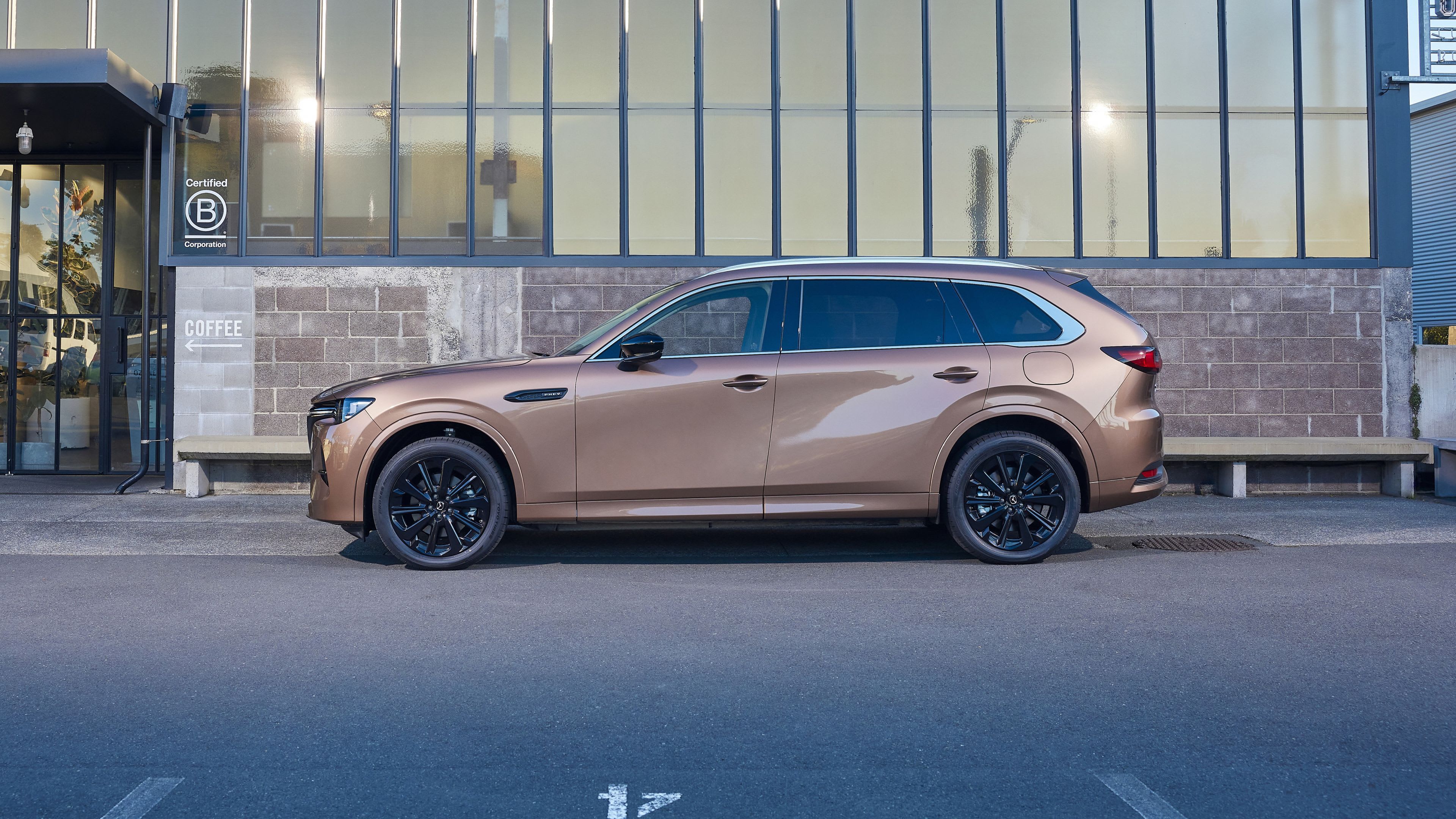 Side view of a Melting Copper Mazda CX-80 7-seat hybrid SUV parked in front of a modern glass and brick office building