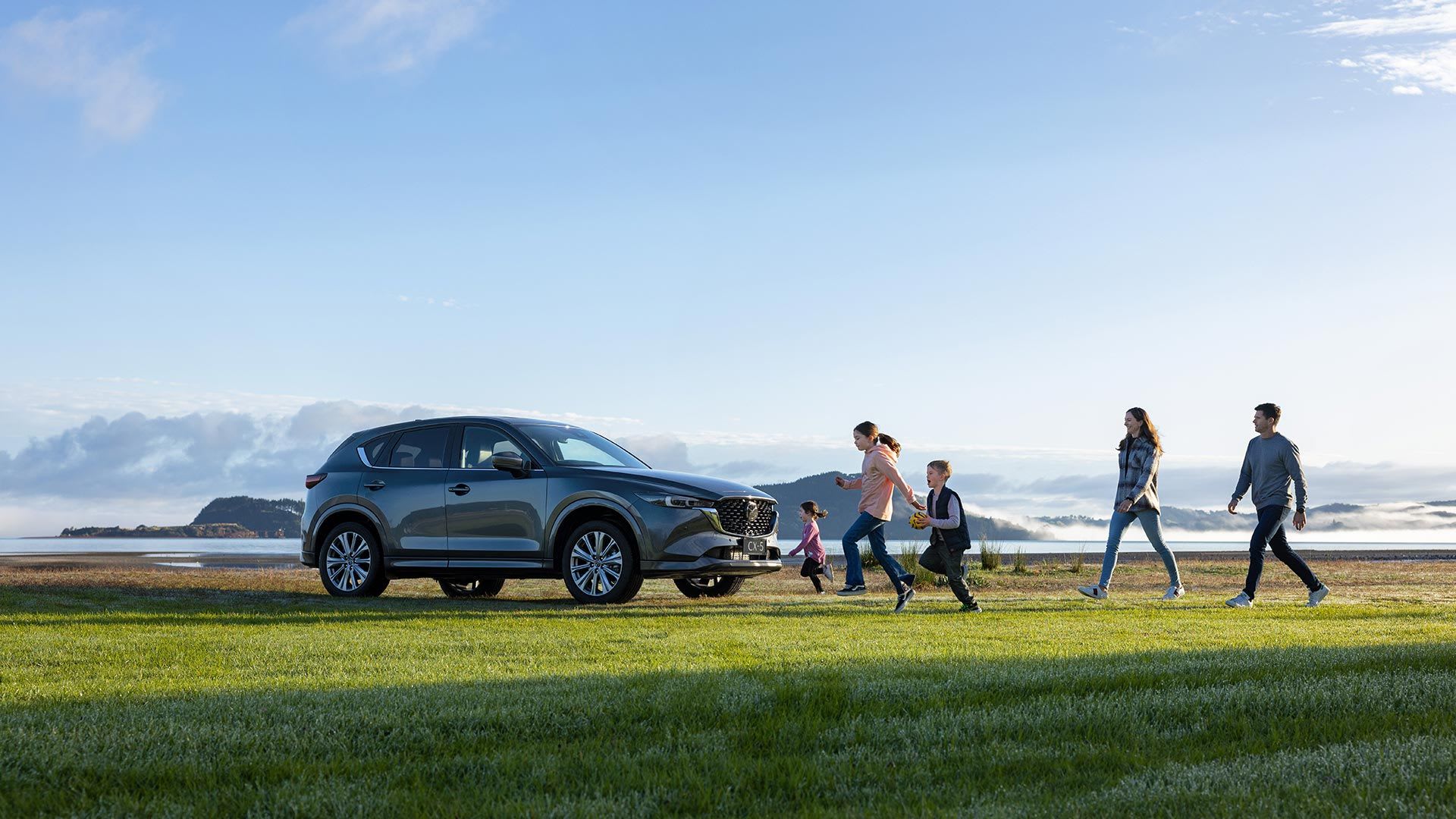 Grey Mazda CX-5 SUV parked on a grass field by a lake with a family of five walking towards the car