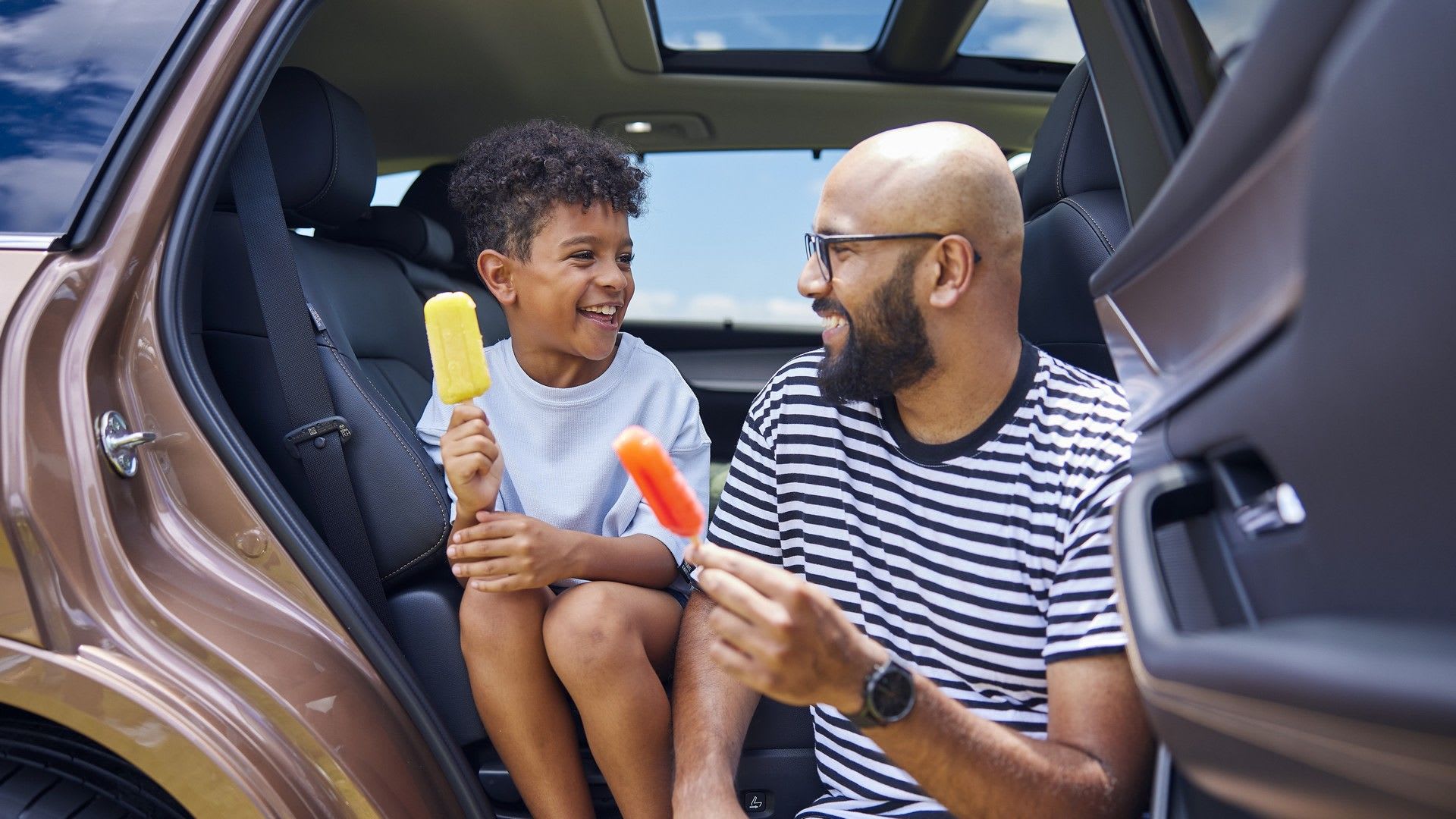 Parent and child enjoying ice lollies while they enjoy the sun in their Mazda CX-80 in Melting Copper Metallic