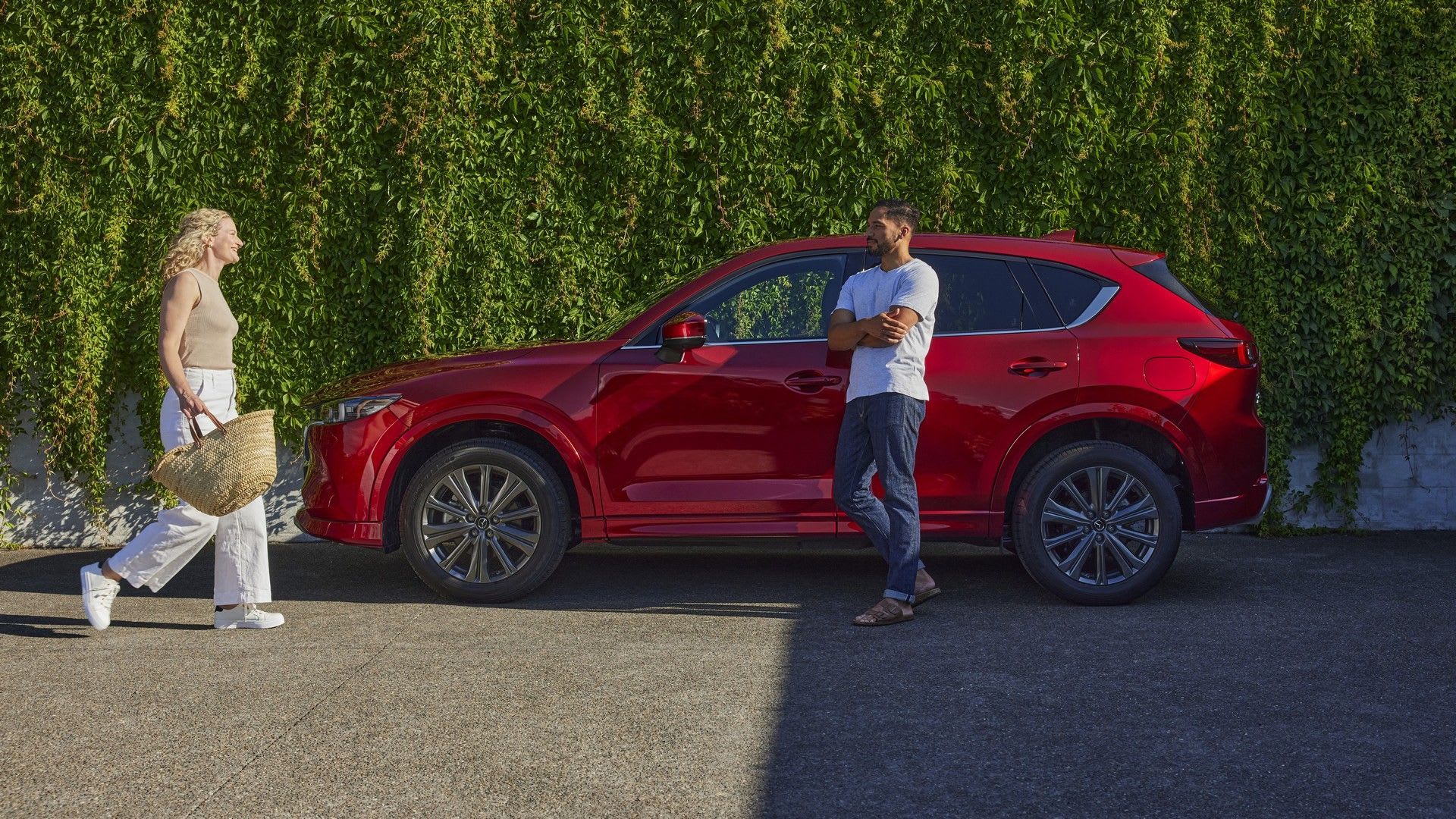 Woman walking towards a man standing next to their Mazda CX-5 SUV in Soul Red Crystal Metallic parked in front of a green hedge 