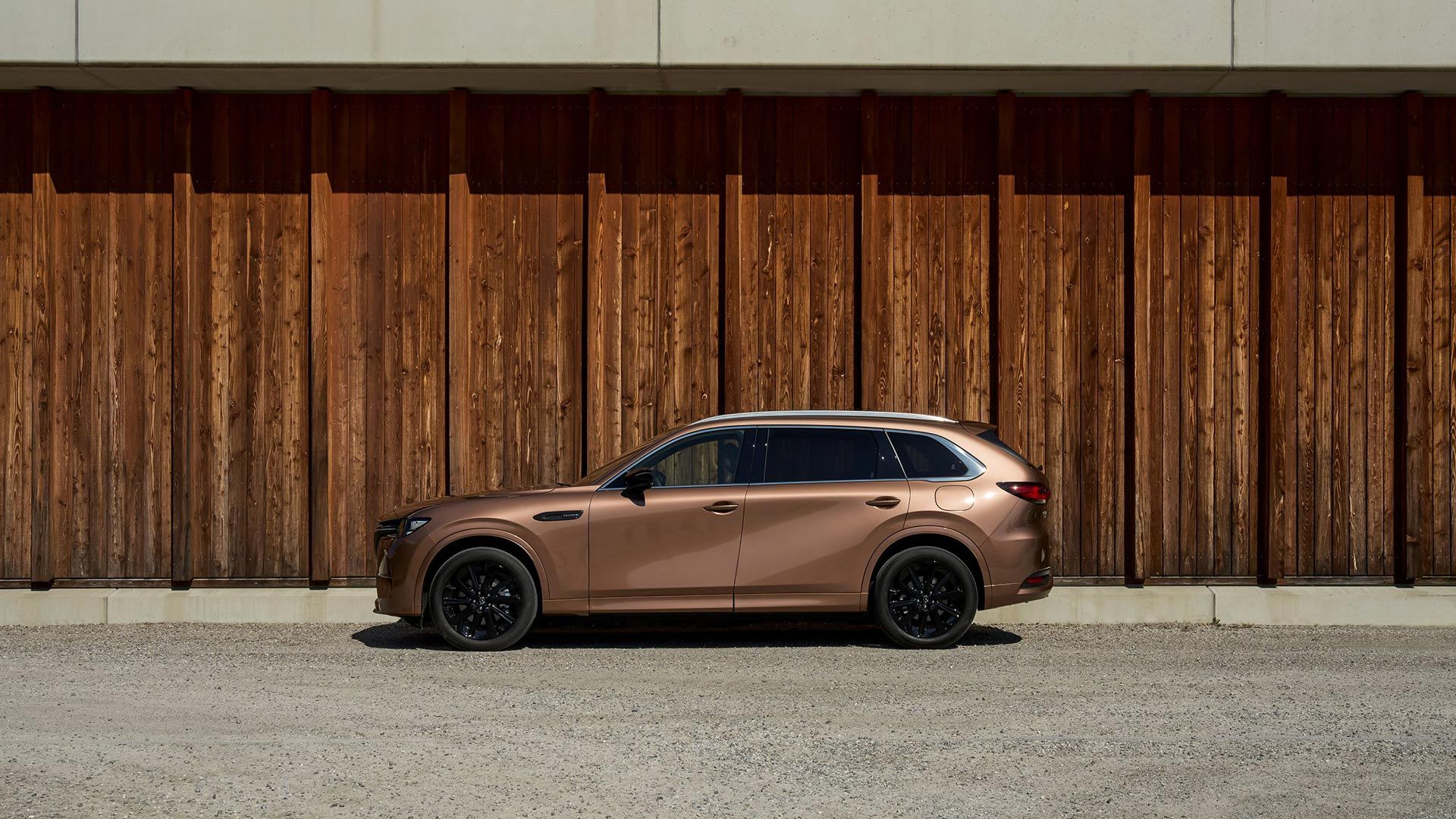 Side view of a Melting Copper Mazda CX-80 SUV parked in front of a wooden wall 