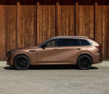 Side view of a Melting Copper Mazda CX-80 SUV parked in front of a wooden wall 