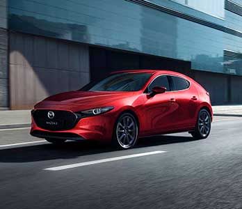 Front three quarter view of a Soul Red Mazda3 driving past a glass city building