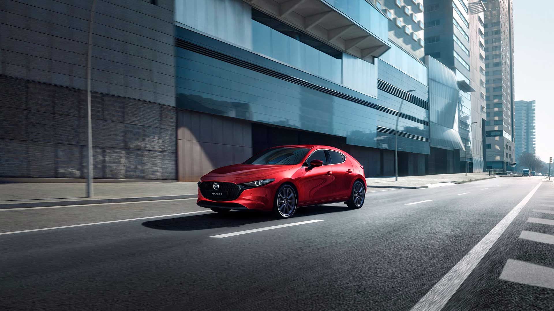 Front three quarter view of a Soul Red Mazda3 driving past a glass city building