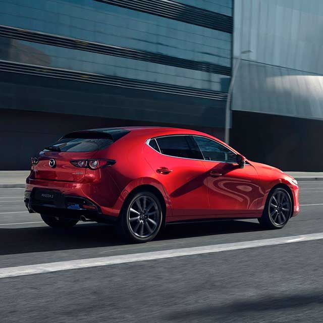 Rear three quarter view of a Soul Red Mazda3 hatchback driving in the city past a glass building