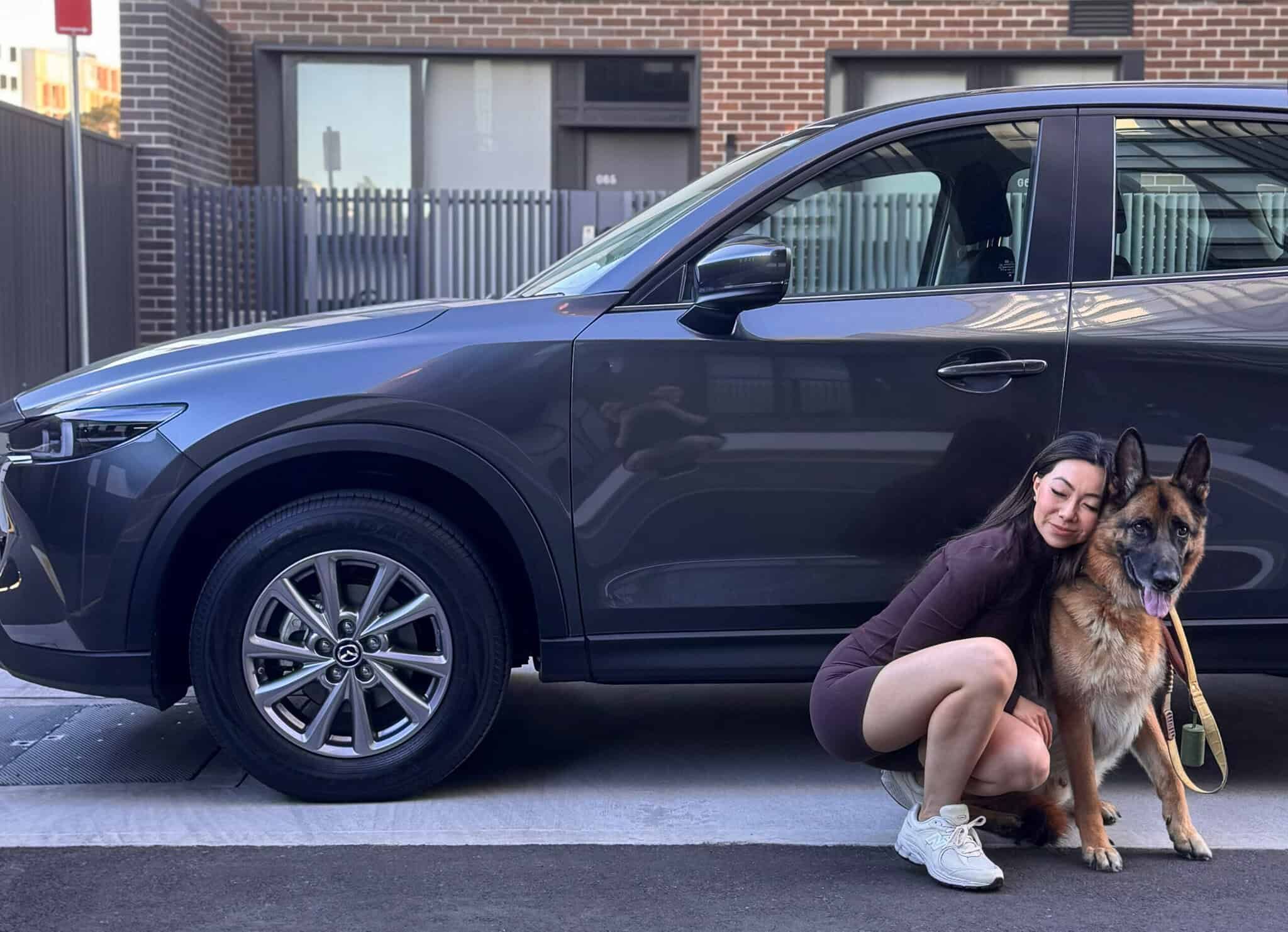 Woman with her dog crouching next to her grey Mazda