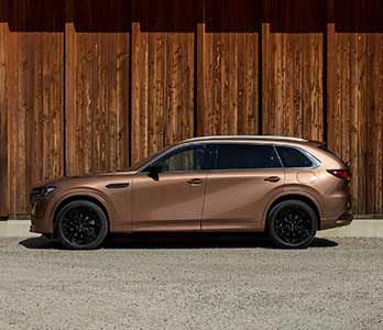 Side view of a Melting Copper Mazda CX-80 SUV parked in front of a wooden wall 