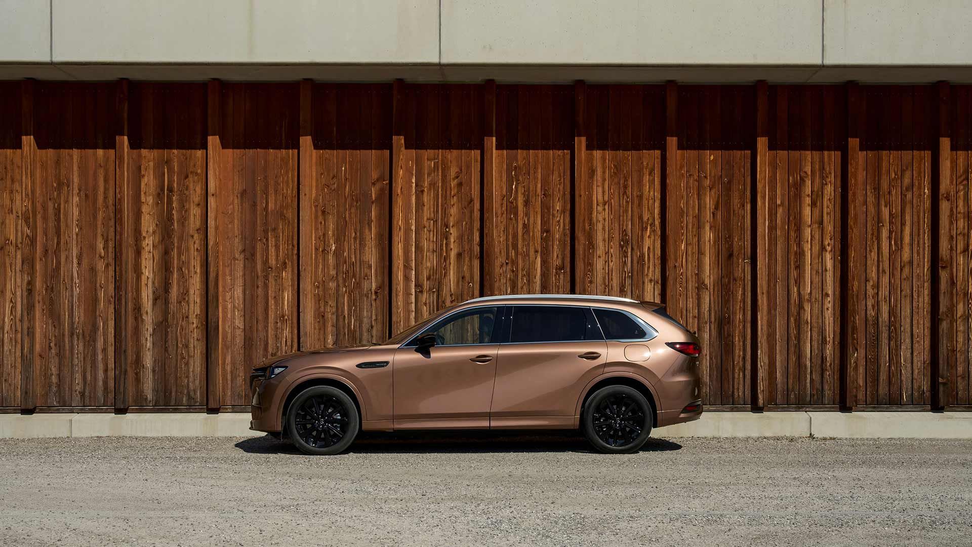 Side view of a Melting Copper Mazda CX-80 SUV parked in front of a wooden wall