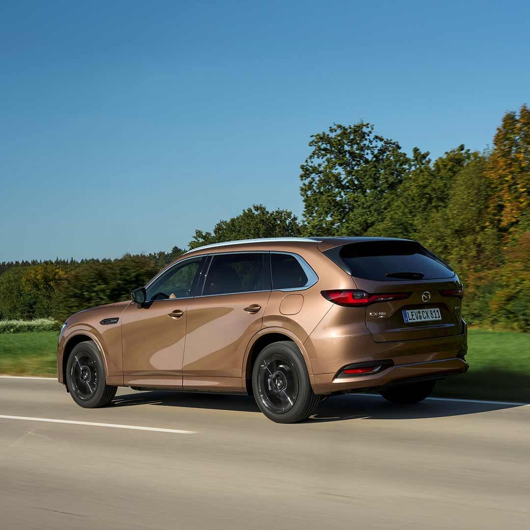 Rear three quarter view of a Melting Copper Mazda CX-80 SUV hybrid driving on the open road