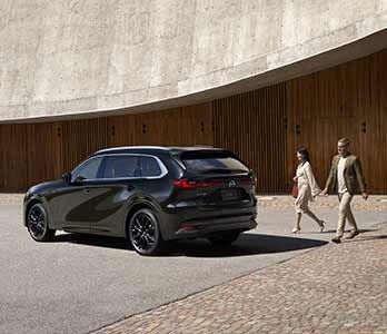 Rear three quarter view of a Jet Black Mazda CX-80 SUV parked in front of a concrete and wood building with a man and woman walking towards the car