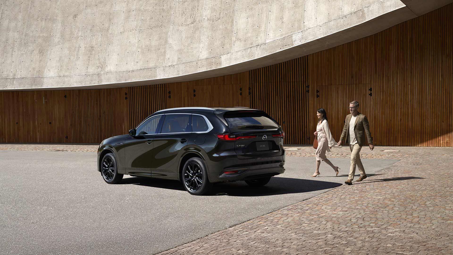 Rear three quarter view of a Jet Black Mazda CX-80 SUV parked in front of a concrete and wood building with a man and woman walking towards the car