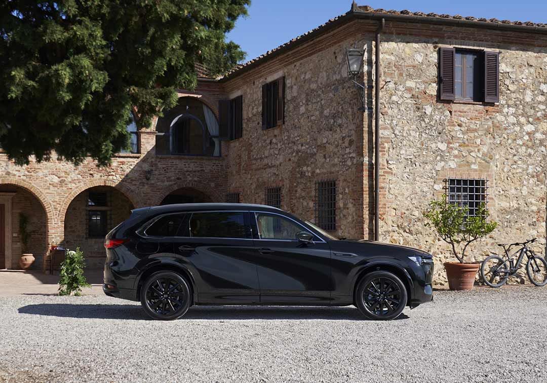Side view of a Jet Black Mazda CX-80 SUV parked in front of a stone Tuscan-style house 