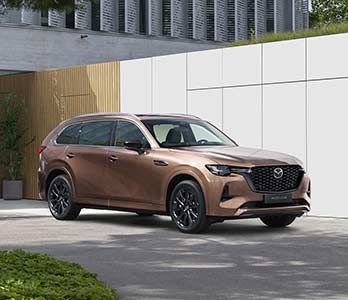 Front three quarter view of a Melting Copper Mazda CX-80 SUV parked in front of a designer house