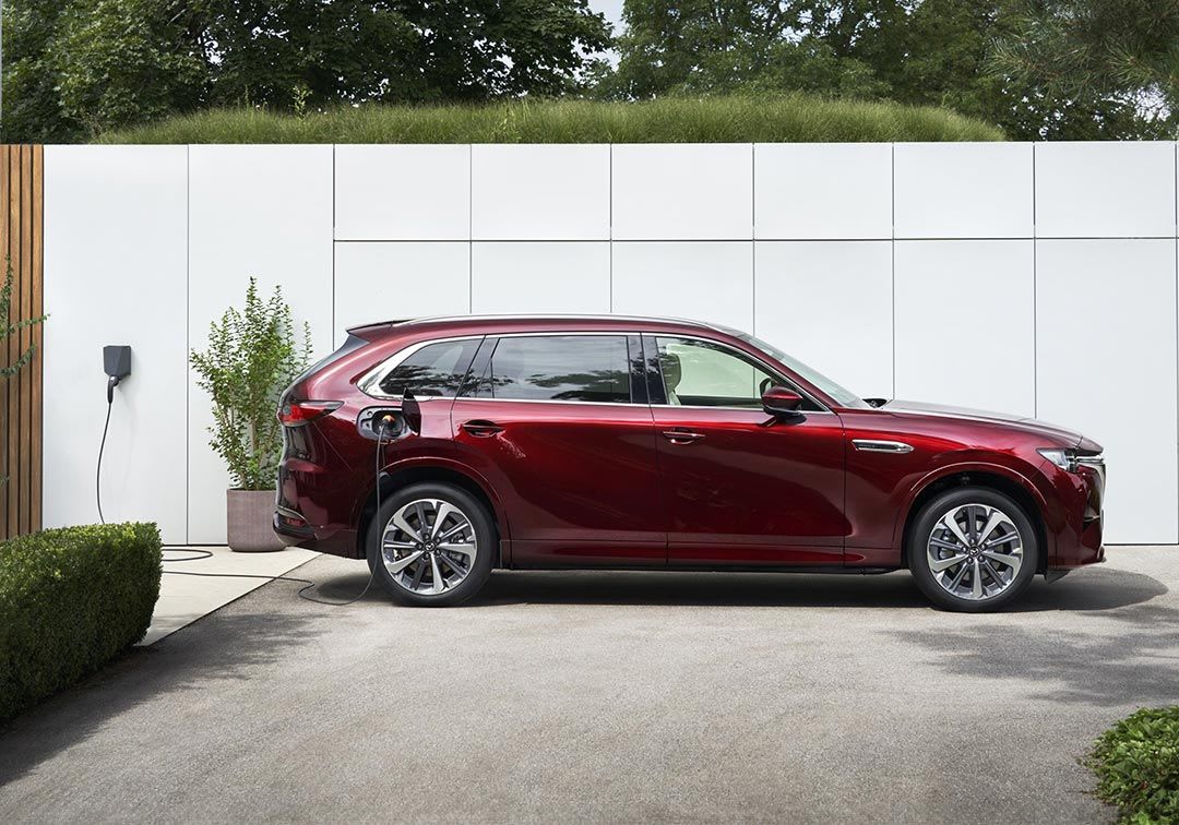 Side view of an Artisan Red Mazda CX-80 plugged in and charging in a driveway