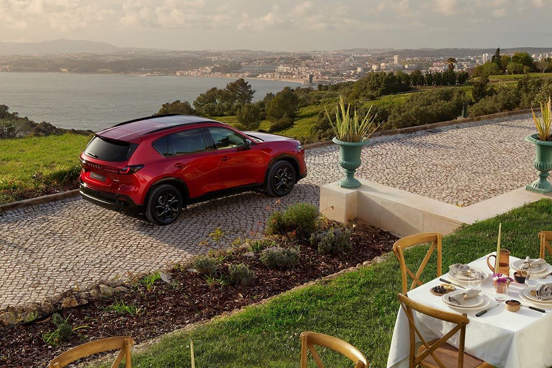High rear three quarter view of a red all-new Mazda CX-5 SUV parked on a stone driveway with a fancy outdoor table setting seen in the foreground