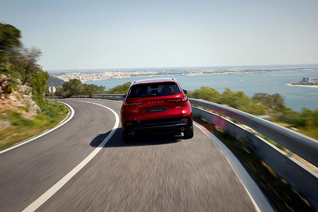 Rear view of a red all0-new Mazda CX-5 SUV driving on a scenic road with the ocean in the background