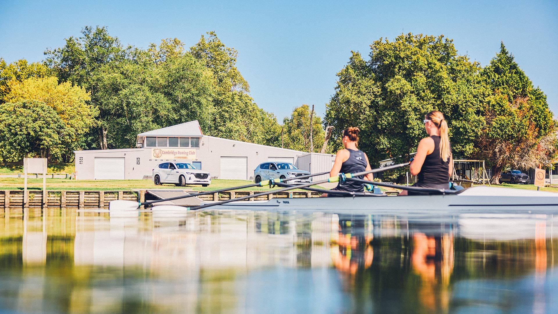 Mazda NZ ambassadors - champion rowers Brooke Francis and Lucy Spoors rowing on a lake with their white Mazda CX-60 SUVs visible on the shore in the background