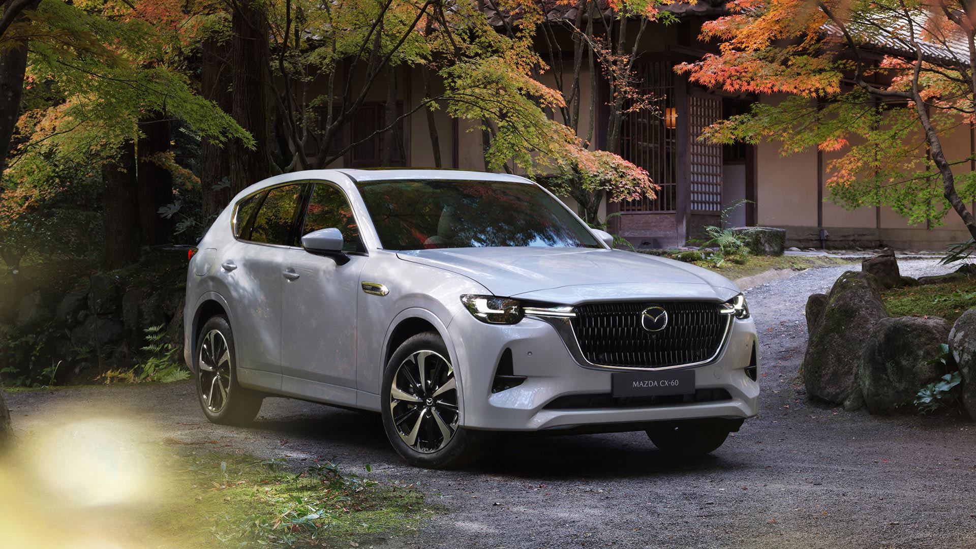 Front three quarter view of a Rhodium White Mazda CX-60 SUV parked on a driveway surrounded by autumnal trees
