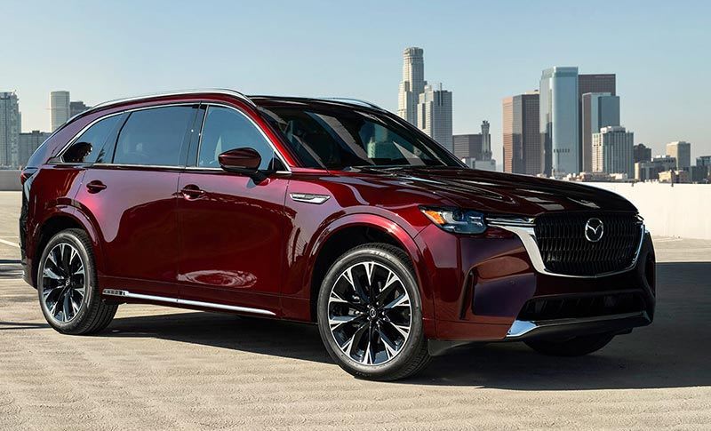 Front three quarter view of an Artisan Red Mazda CX-90 SUV in a carpark with a cityscape in the background