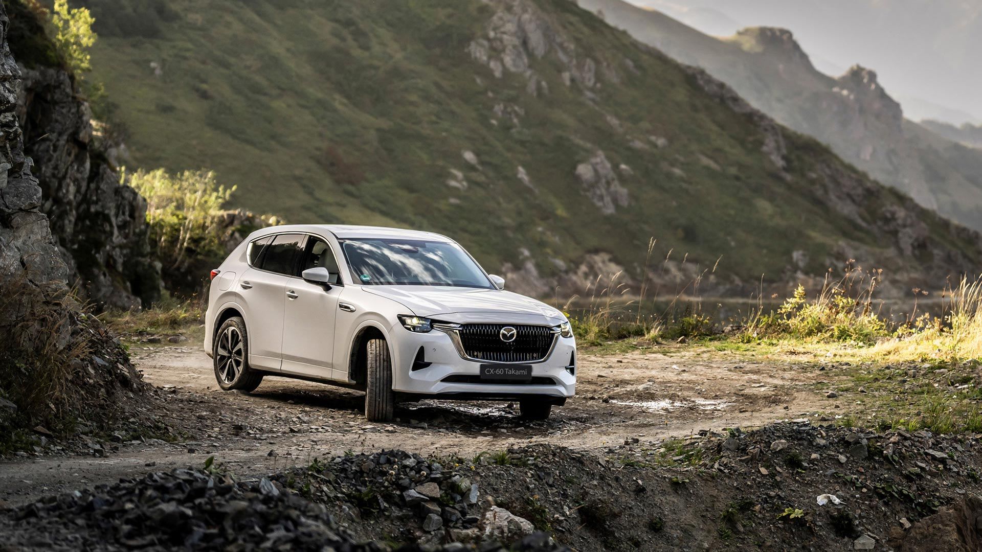 Front three quarter view of a Rhodium White Mazda CX-60 SUV driving on an unpaved mountain road
