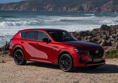 Front three quarter view of an Artisan Mazda CX-60 SUV parked in an unpaved carpark in front of a beach