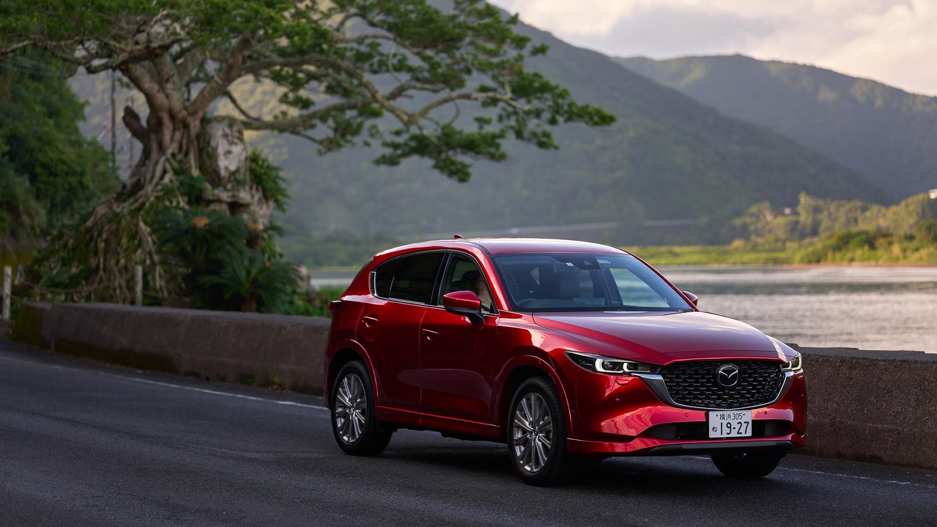 Front three quarter view of a Soul Red Mazda CX-5 SUV driving next to a lake in Japan