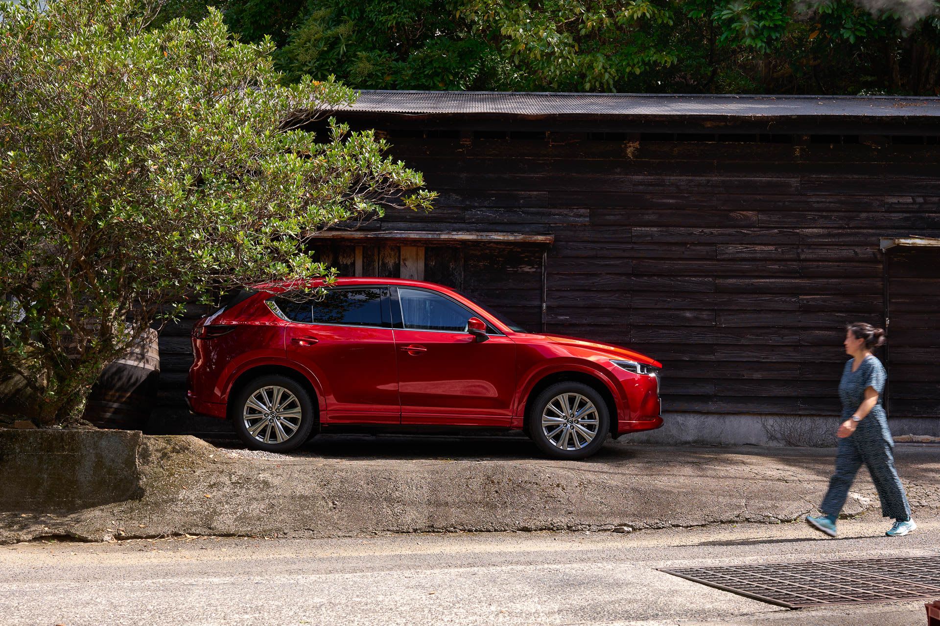 Side view of a Soul Red Mazda CX-5 SUV parked in front of a house in Japan's Amami Oshima, with a woman walking nearby