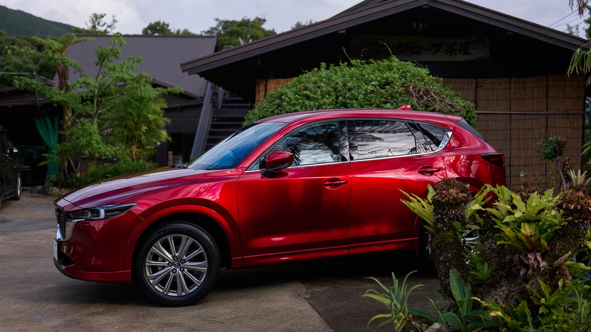 Front three eighths view of a Soul Red Mazda CX-5 SUV parked in front of a wooden house in Japan's Amami Oshima region