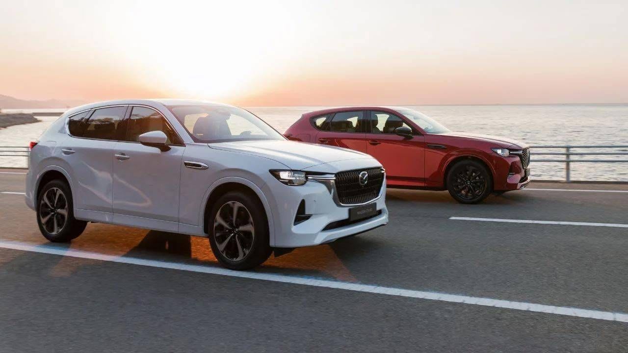 Rhodium White Metallic and Soul Red Crystal Metallic Mazda CX-60 SUV plug-in hybrids driving together on the open road next to the ocean 