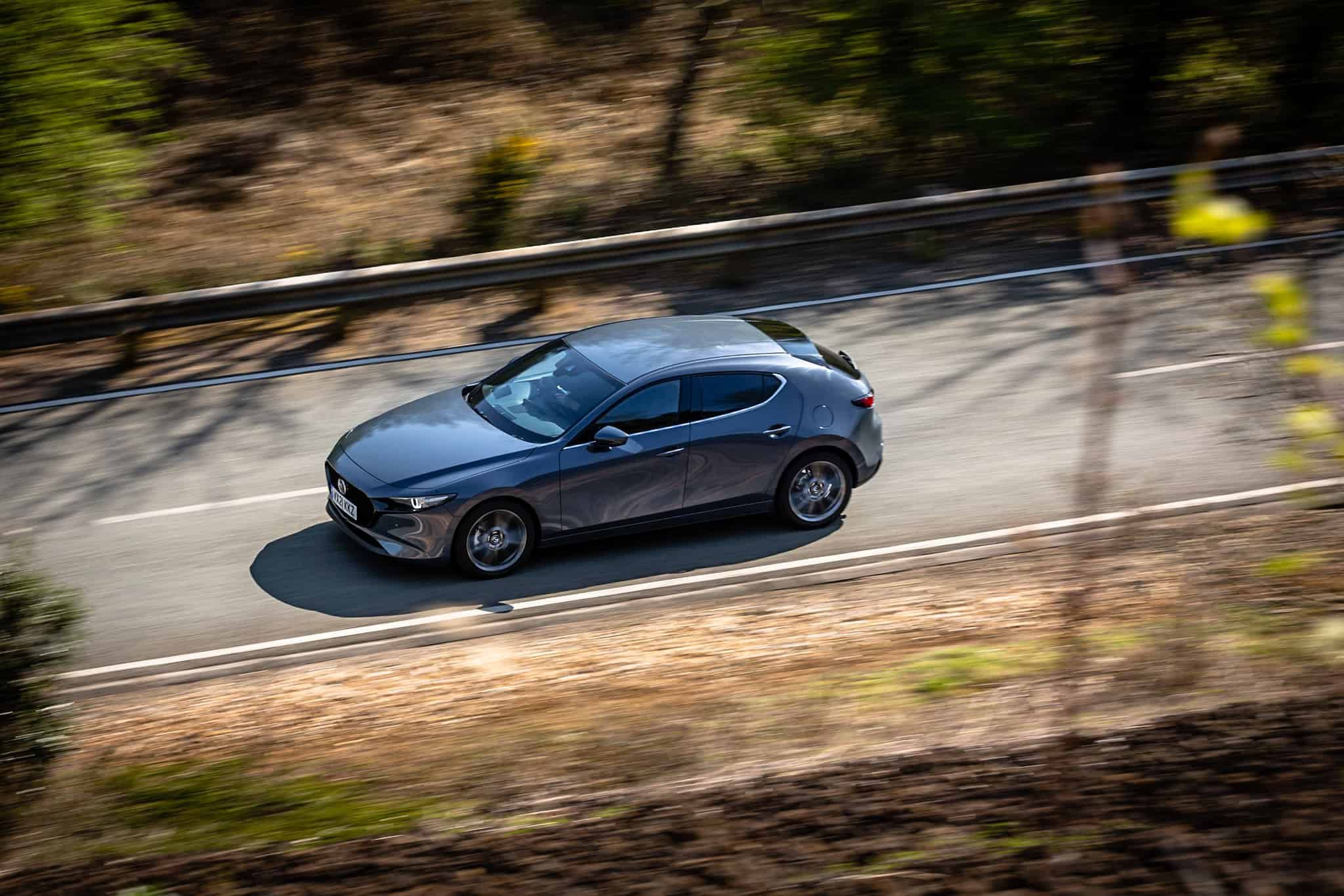 Top down view of a dark grey Mazda3 driving along a road in the woods
