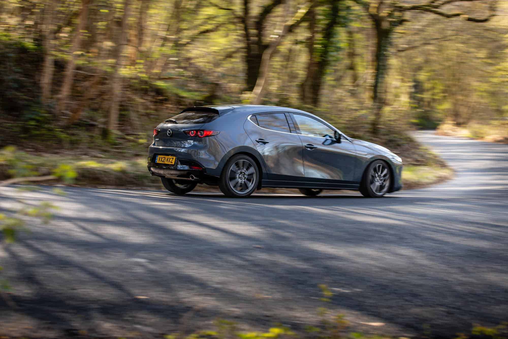 Rear three quarter view of a dark grey Mazda3 driving through a hairpin bend in the woods 