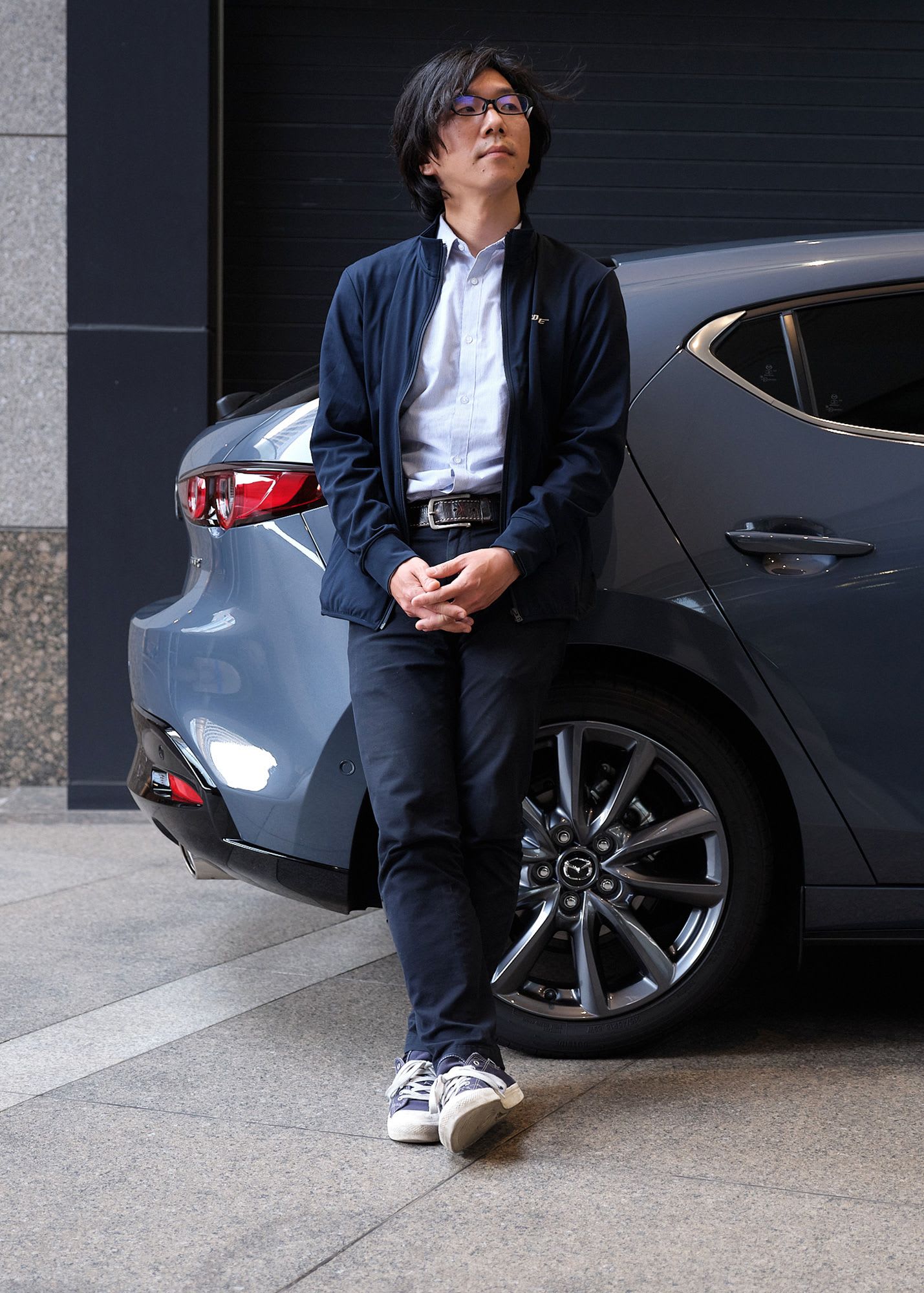 Portrait of Koji Wakamatsu, assistant manager in Mazda’s Electrical & Electronics Performance Development Department leaning against a blue Mazda car