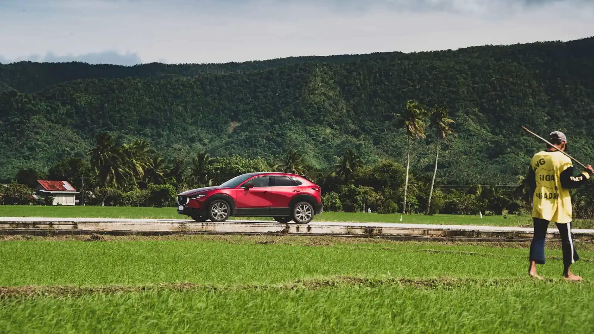 Side view of a Soul Red Mazda CX-30 SUV driving through a filed next to lush mountains in the Philippines