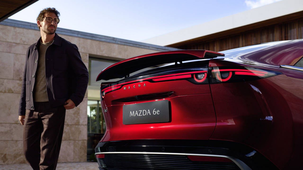 Exterior shot of the Mazda 6e in Soul Red Crystal Metallic, showing the extended rear spoiler as a person walks around the back of the vehicle.