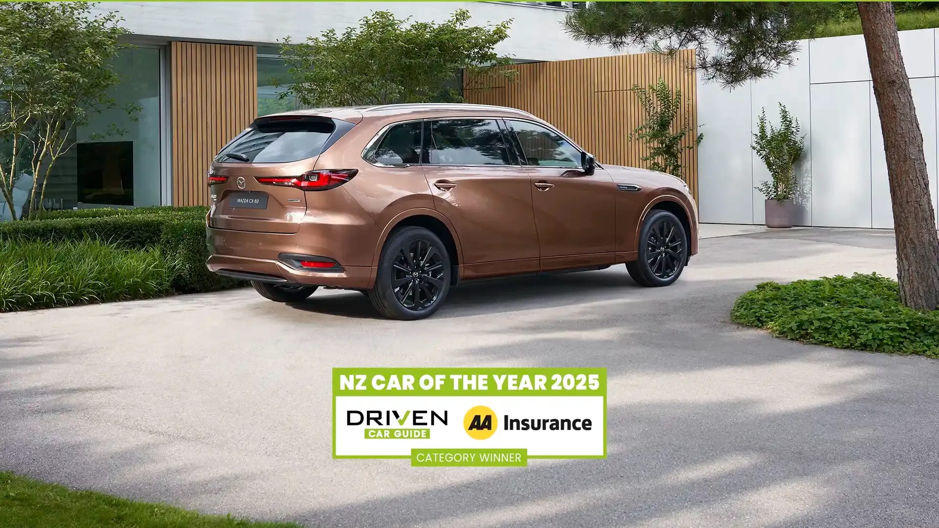 Rear three quarter view of a Melting Copper Mazda CX-80 SUV parked in the driveway of a luxury house