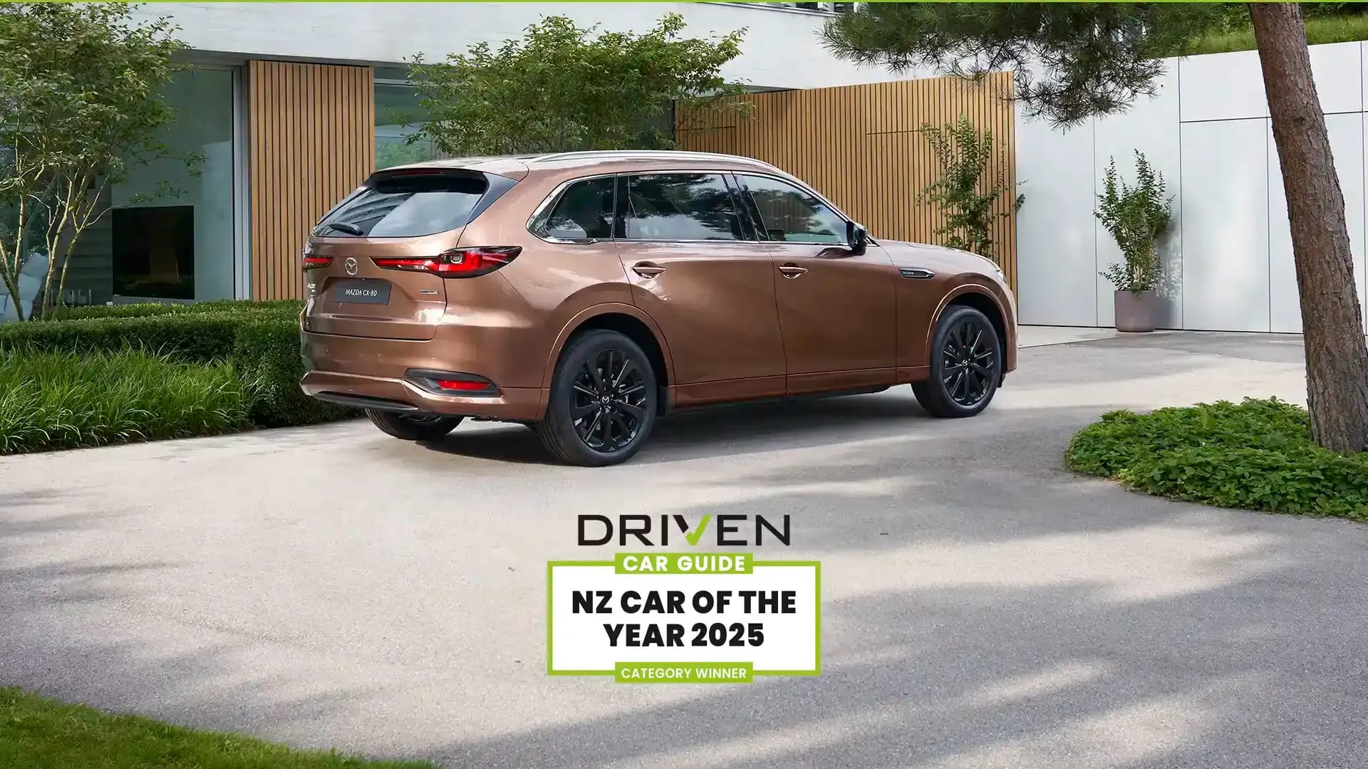 Rear three quarter view of a Melting Copper Mazda CX-80 SUV parked in the driveway of a luxury house